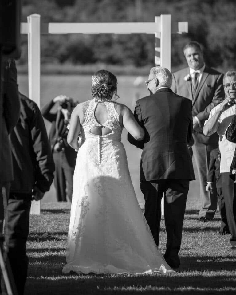 Outdoor wedding scene: bride in a lace gown walking arm-in-arm with a man in a suit toward a white ceremony arch (black-and-white photo).