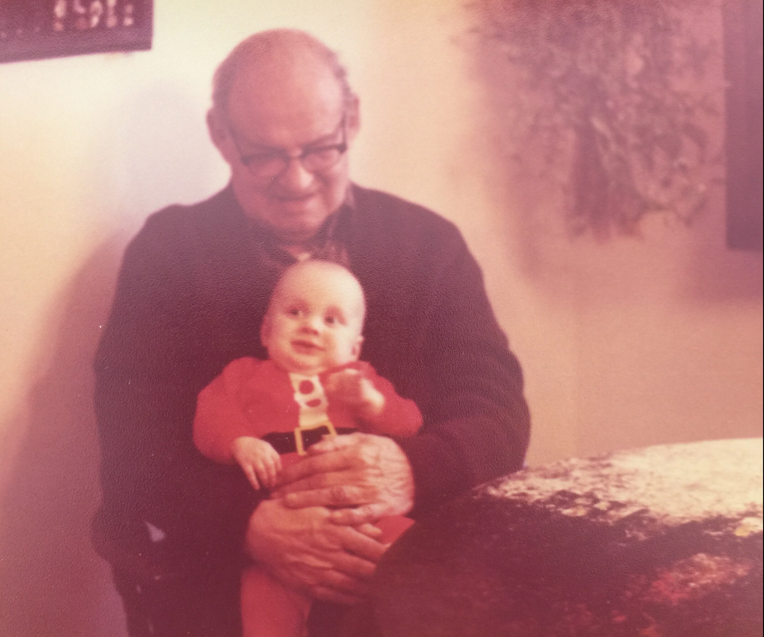 Elderly man with glasses sitting indoors, holding a baby doll on his lap.