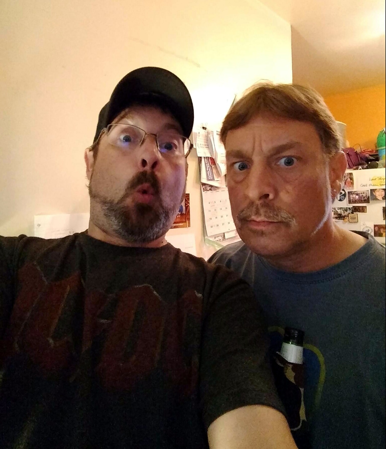 Two men posing for a goofy selfie indoors, one wearing a cap, both with surprised, wide-eyed expressions.