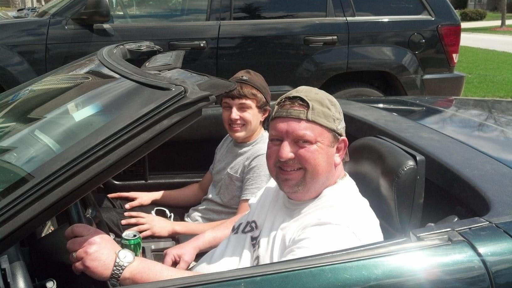 Two men in a green convertible with the top down, smiling at the camera, wearing casual shirts and caps.