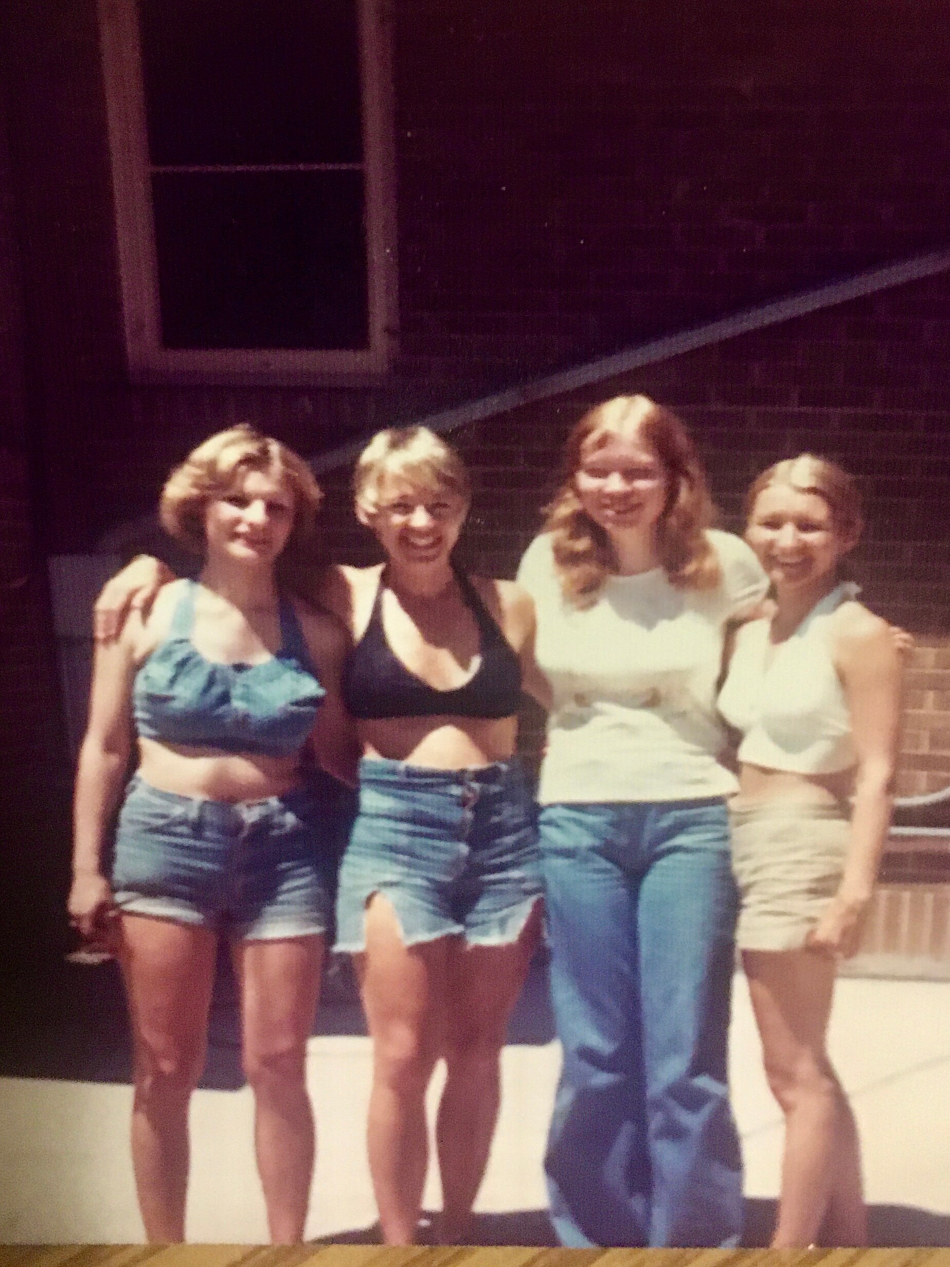 Five young women posing together outdoors, smiling, in casual summer outfits with denim shorts and tank tops forgetfully close together in front of a brick backdrop.