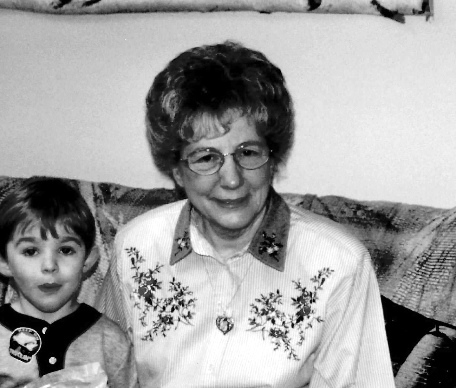 Older woman with short hair and glasses sits on a patterned couch, wearing a light striped blouse with floral embroidery, next to a young girl partially visible on the left.