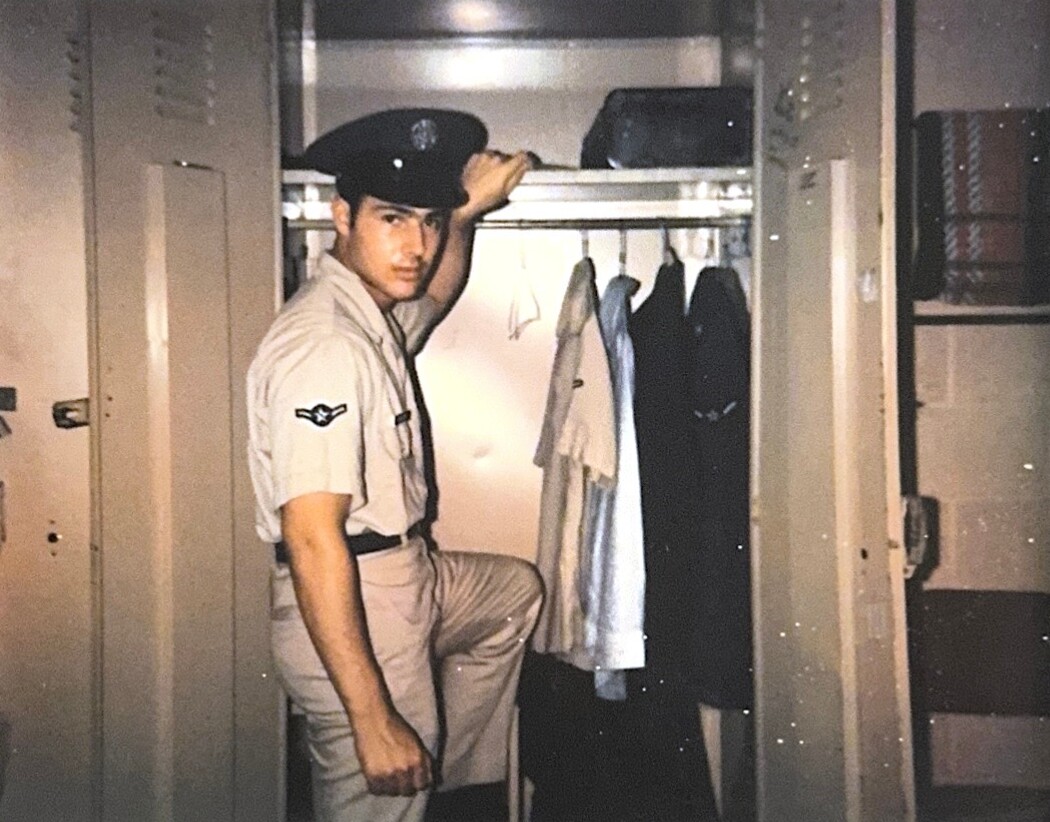 Uniformed man in a beige military-style outfit stands inside an open locker, one knee on the shelf, looking at the camera.
