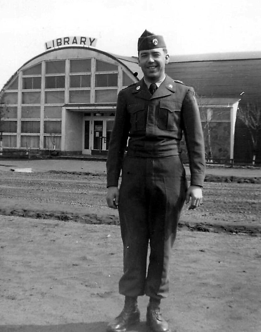 Man in a vintage military uniform standing outside a library with a glass-front entrance, black-and-white photo.