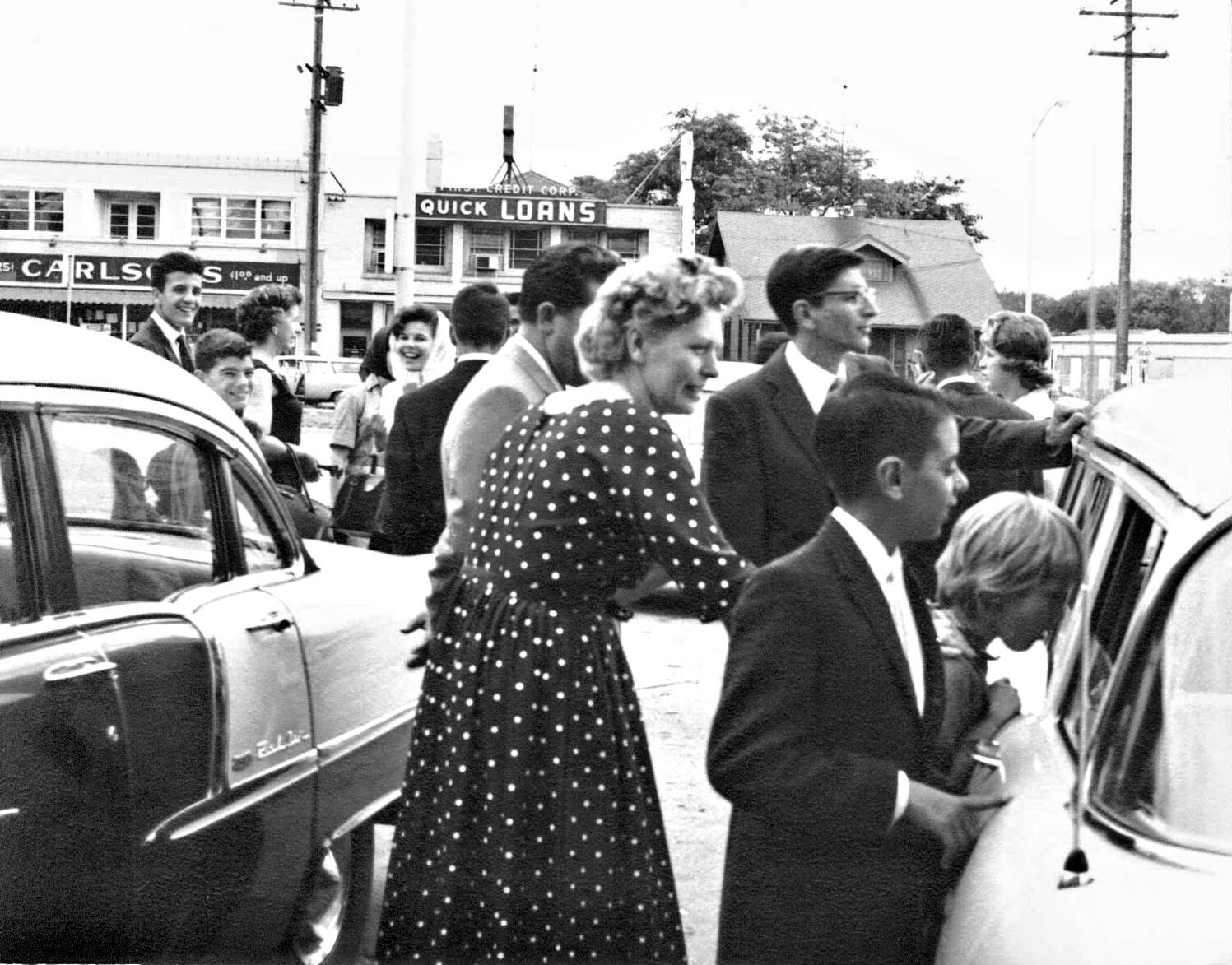 Crowd of people in 1950s attire around vintage cars on a street; a woman in a polka-dot dress steps toward a car with others watching nearby.