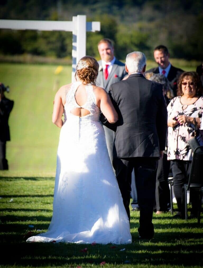 Bride in a white wedding gown walking arm-in-arm with an older man down the outdoor ceremony aisle, guests watching in the background.
