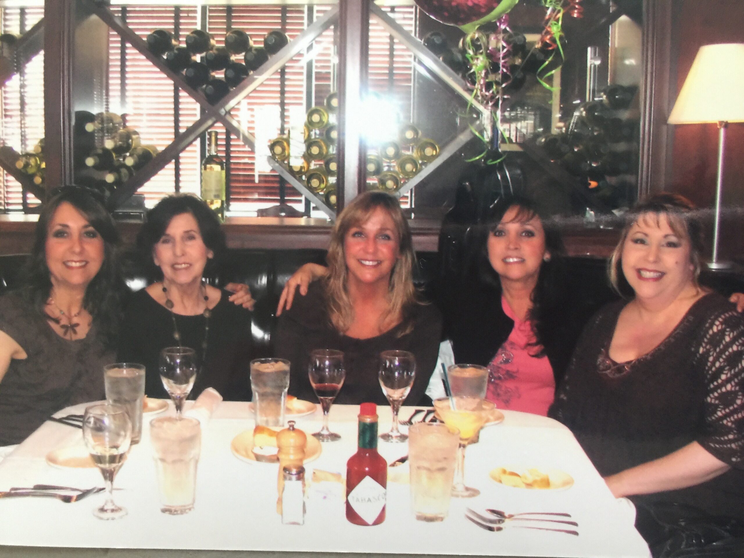 Five women seated at a restaurant table, smiling, with drinks and plates; wine bottles on a rack behind them.