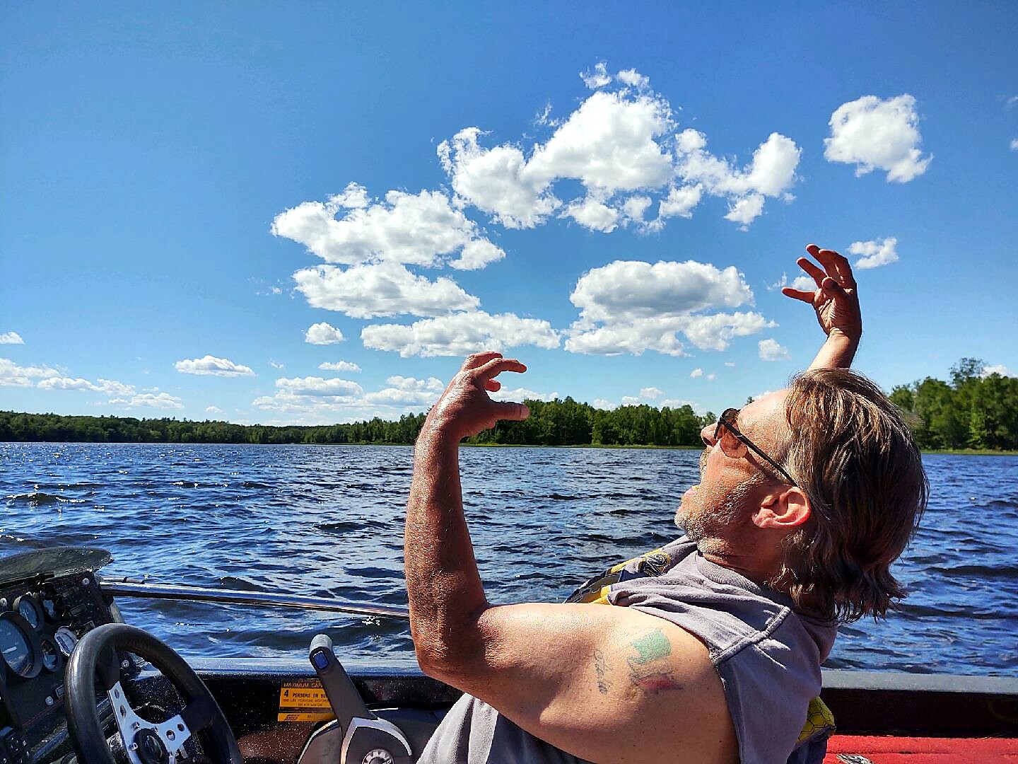 Man wearing sunglasses on a boat with arms raised, enjoying a sunny lake under a blue sky with white clouds.