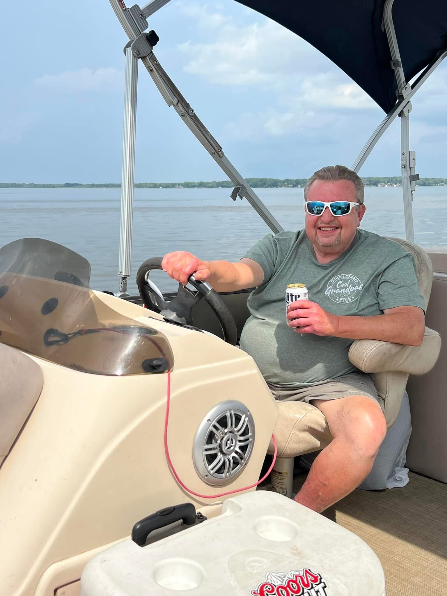 Smiling man wearing sunglasses sits at the boat's helm, holding a beer can with water in the background under a canopy.