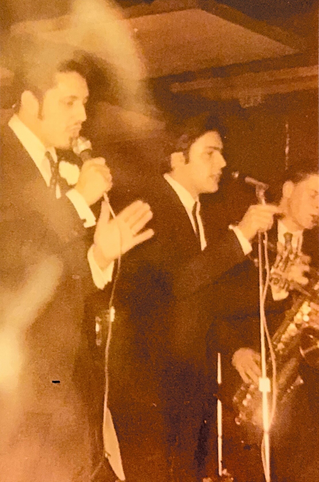 Three men in tuxedos standing at a microphone on a stage, speaking to an audience at a formal event. One man gestures with his hand as he speaks. The image has a vintage sepia tone with floral decor in the foreground.