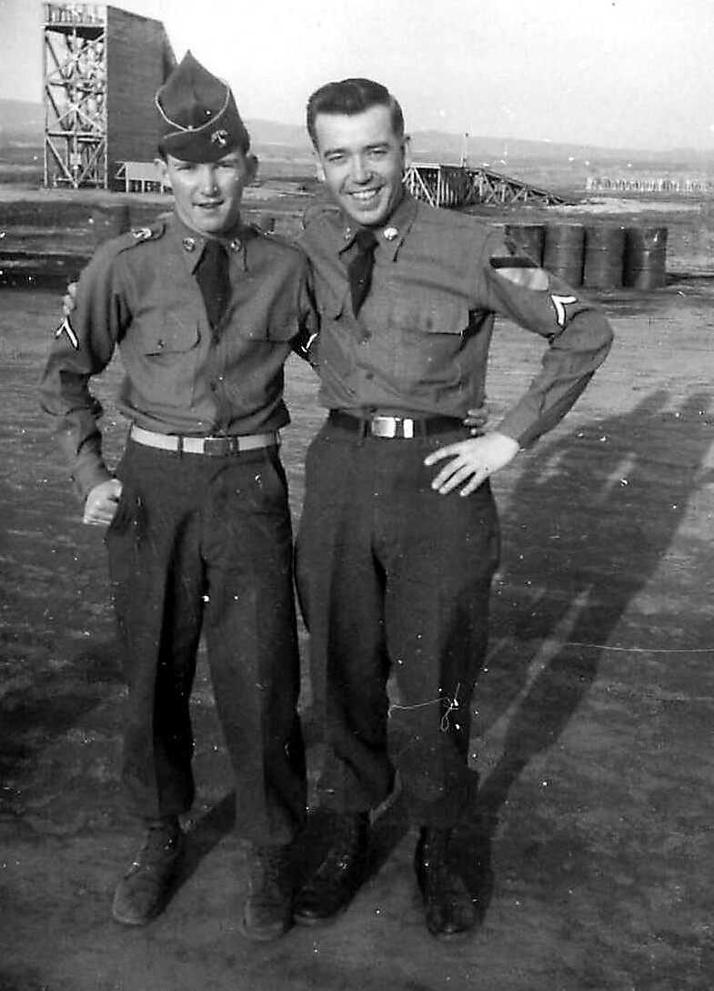 Two young soldiers in uniform posing with arms around each other on a dusty outdoor training ground with structures in the background.