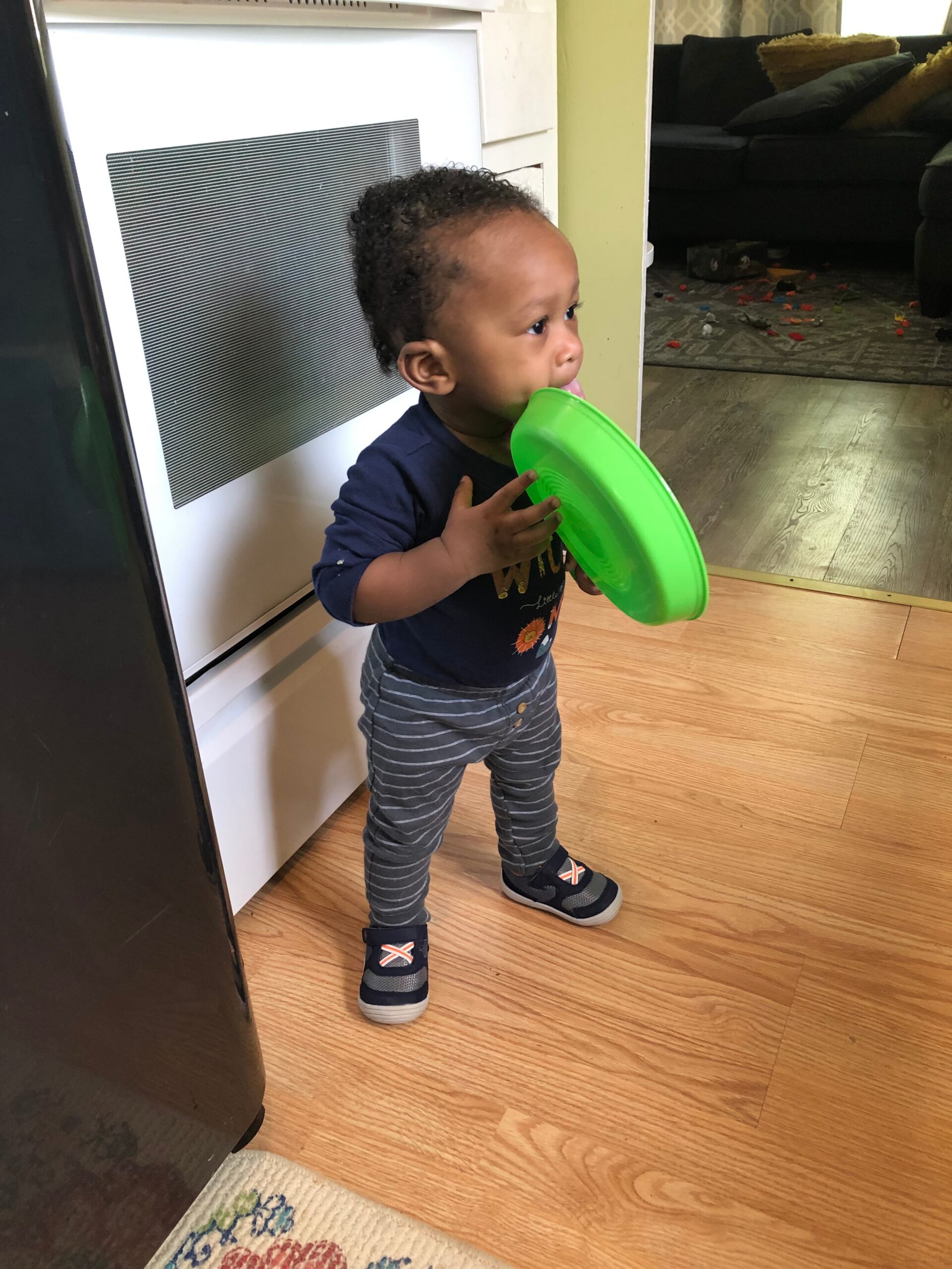 Toddler standing by a kitchen appliance, holding a bright green plate to their mouth as if eating; wearing a navy shirt and striped pants.