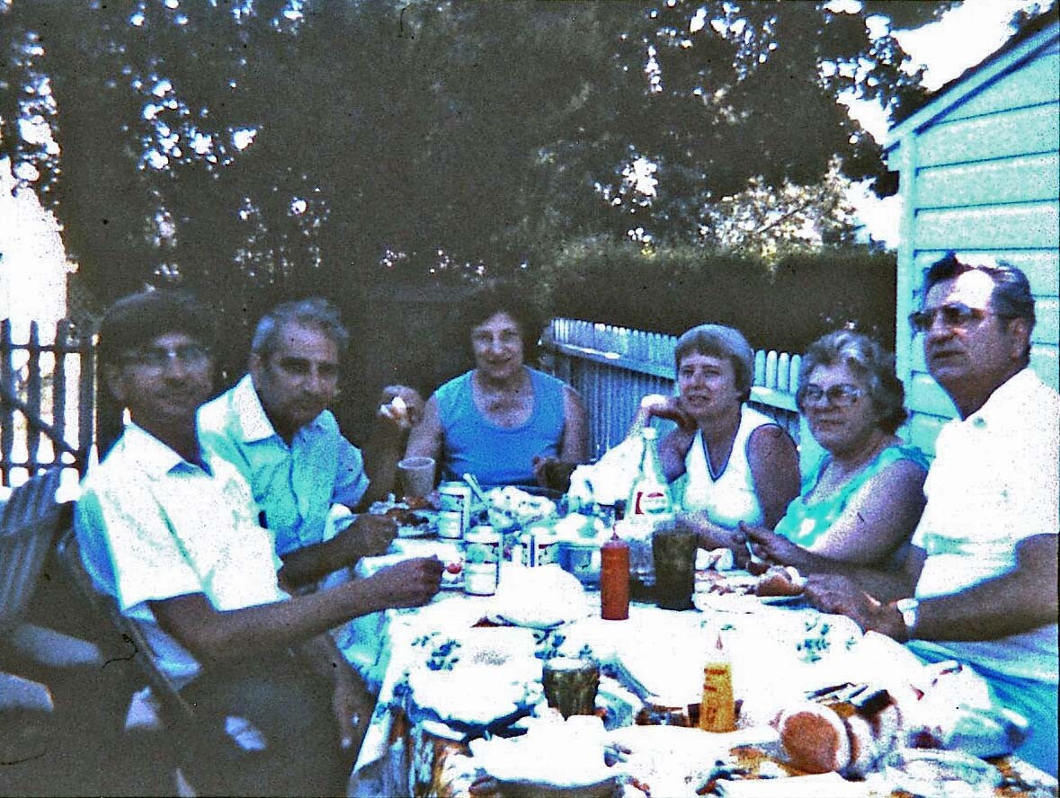 Group of six adults sitting around a picnic table outdoors, enjoying food and drinks.