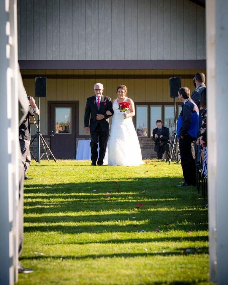 Bride in a white gown holds a red bouquet as she walks arm-in-arm with an older man down a grassy aisle at an outdoor wedding, guests watching on either side.