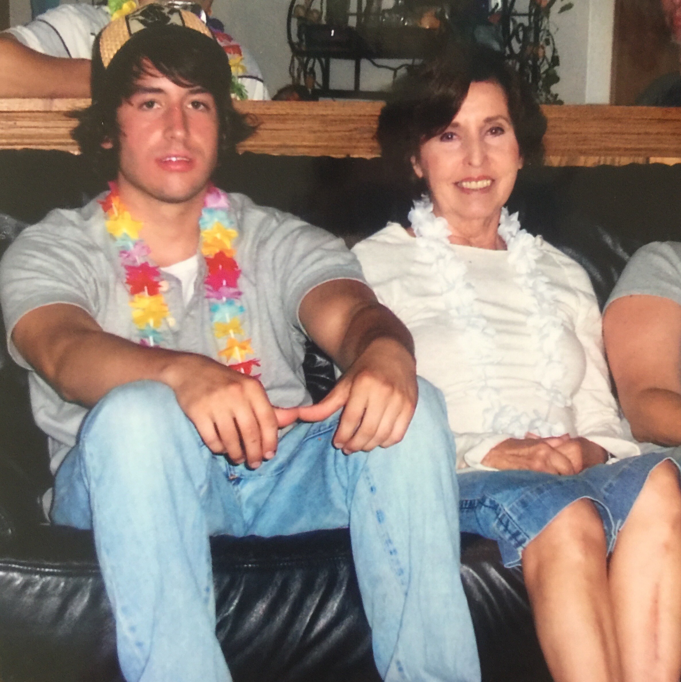 Two people sit close together on a black leather couch wearing colorful Hawaiian leis; smiling and relaxed at a casual gathering.
