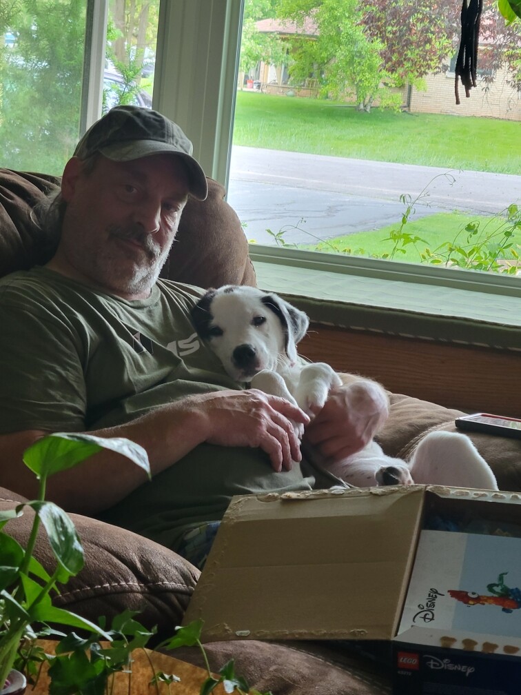 Older man in a green shirt and cap cuddling a small white puppy with black markings on a brown couch by a window.
