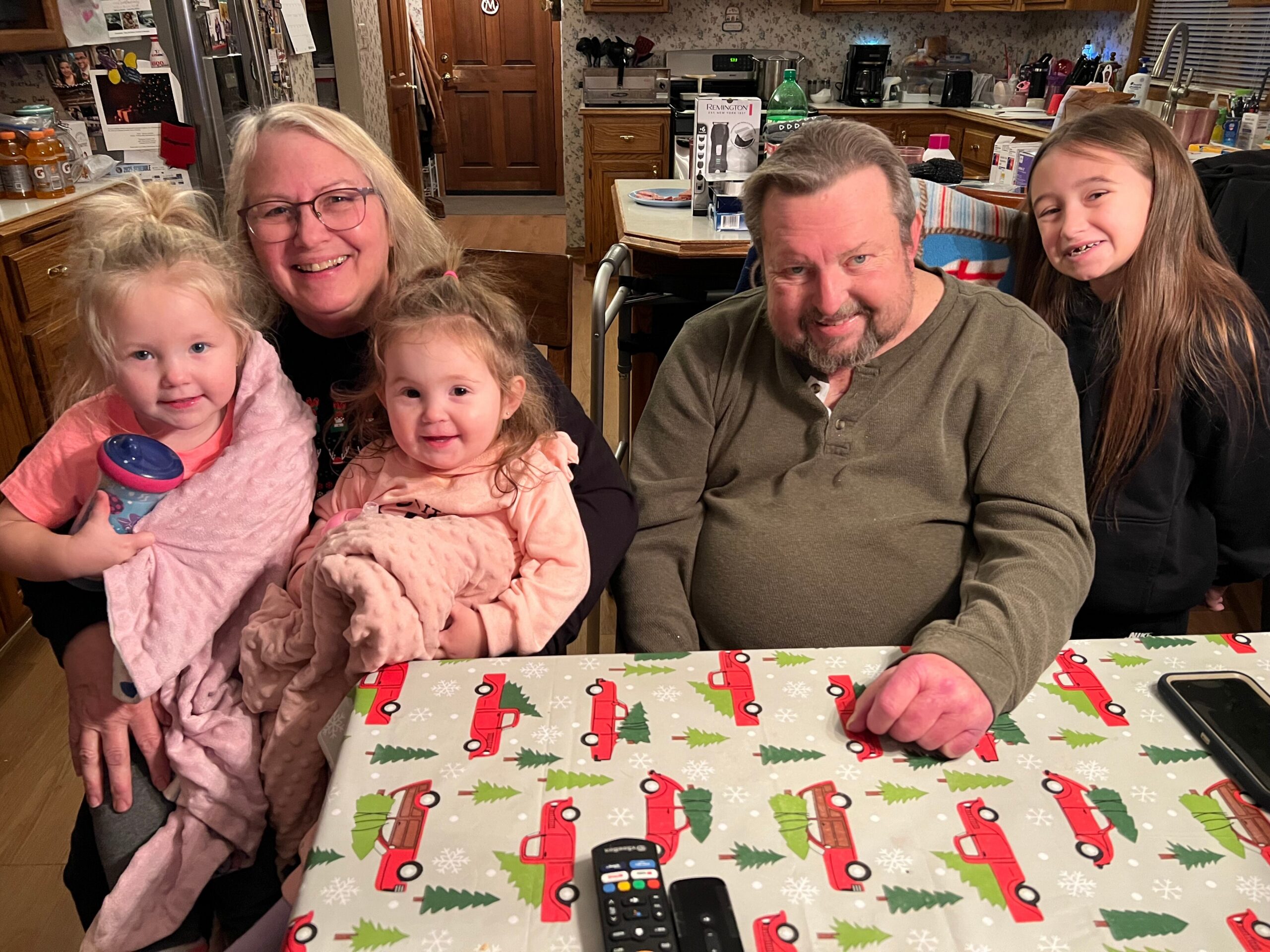 Family group of four with two young girls and two adults posing around a table with holiday gift wrap in a kitchen. Inset items: a remote on the table and a gift box ahead of them.