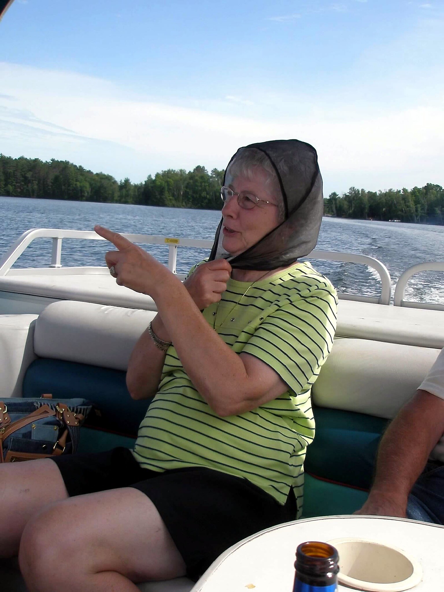 Older woman on a boat pointing to the left, wearing a sheer black hood and lime striped shirt, seated near a drink on deck with water and trees in the background