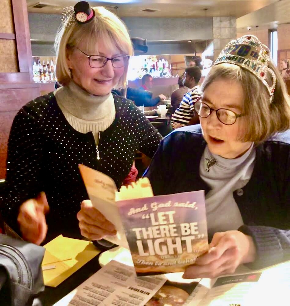 Two smiling women at a restaurant table, one wearing a birthday crown, reading a card that says 'Let there be light'.