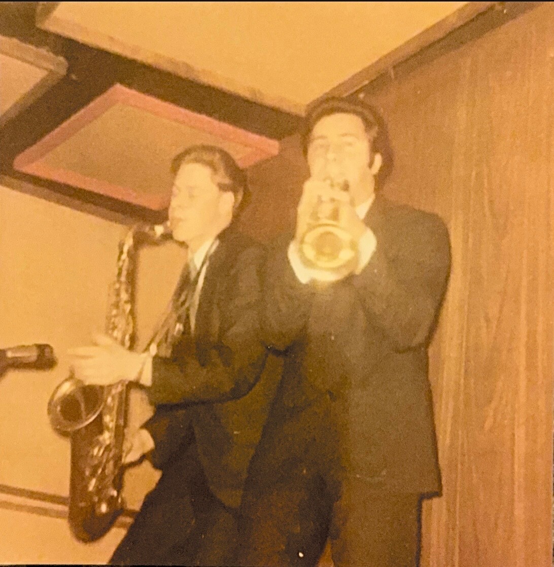 Two men in suits performing live; one plays a saxophone while the other holds a bottle toward the camera.