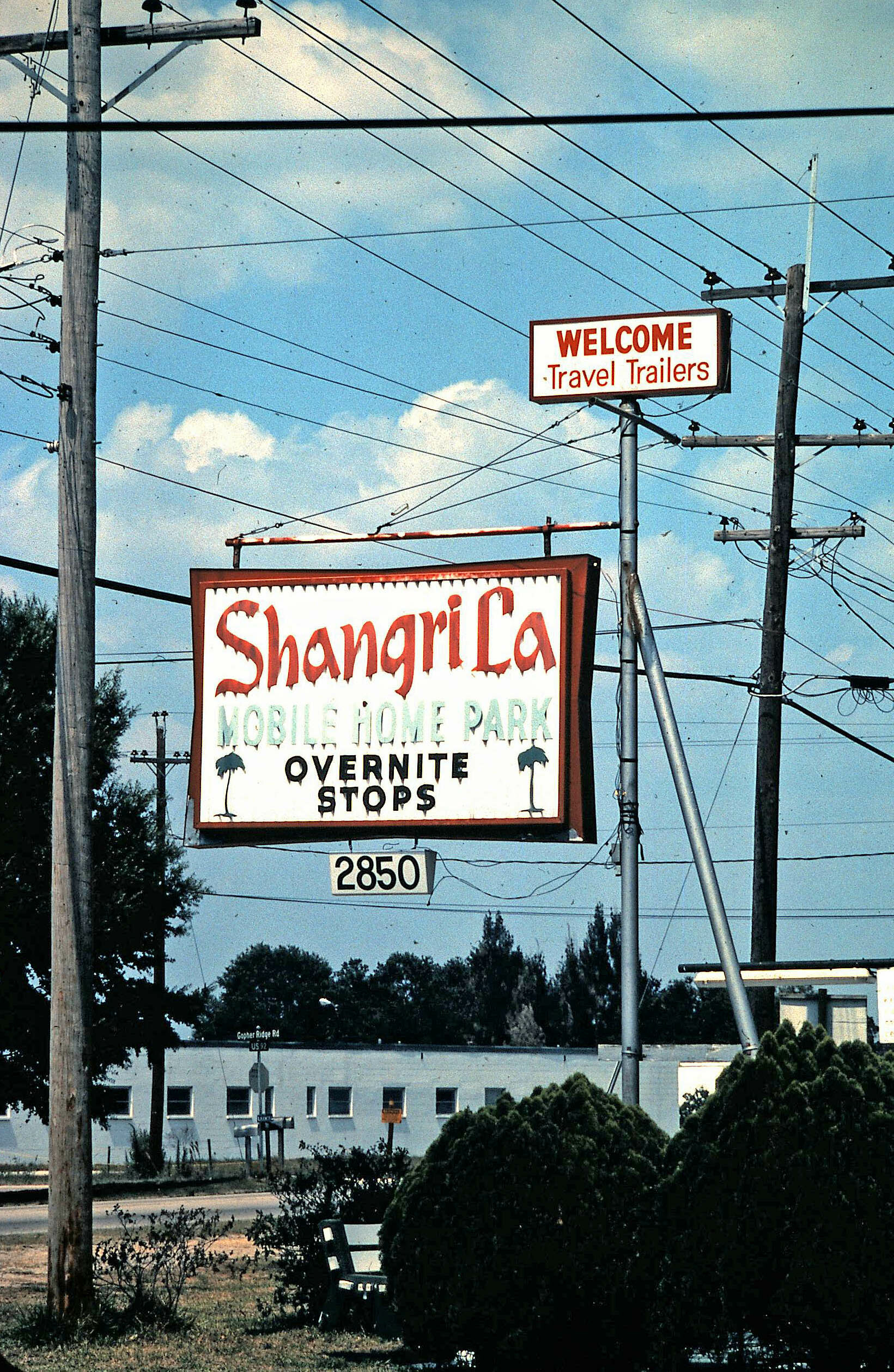 Roadside scene with utility poles and signs reading 'Welcome Travel Trailers' and 'ShangriLa Mobile Home Park' by a street.