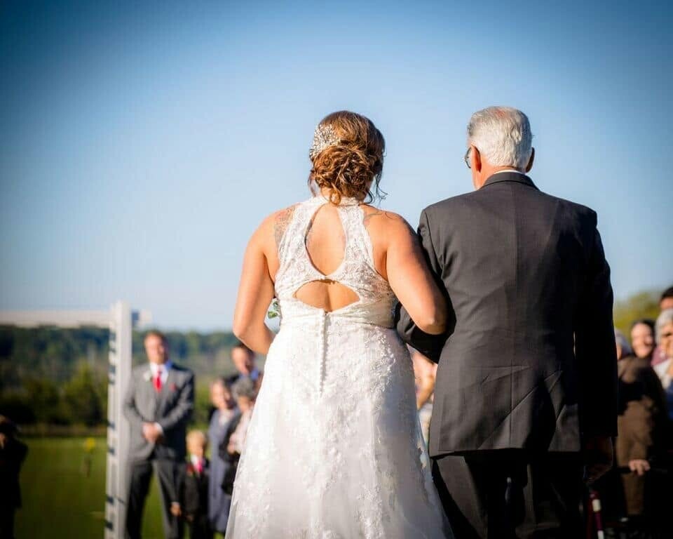 Bride in a lace white wedding gown walking arm-in-arm with an older man in a suit during an outdoor ceremony, guests blurred in the background.