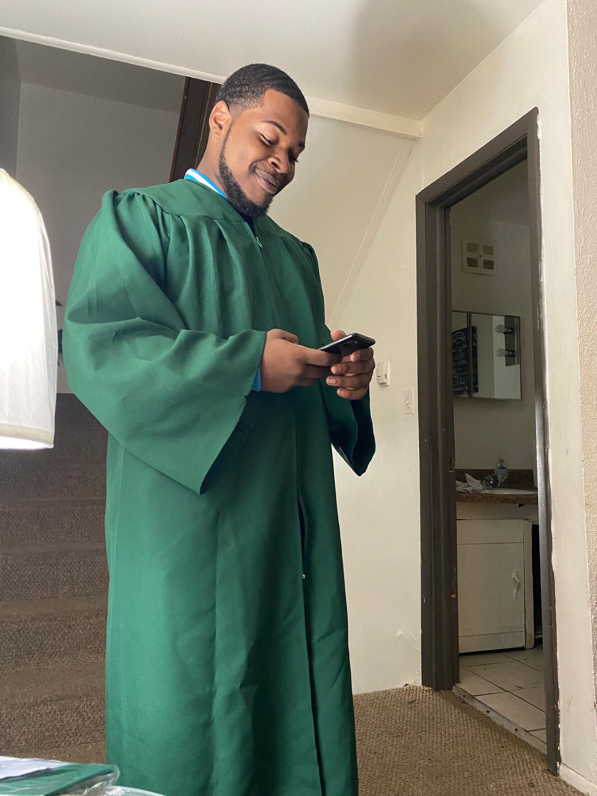 Young man in a green graduation gown looking at his phone indoors with a small smile on his face.