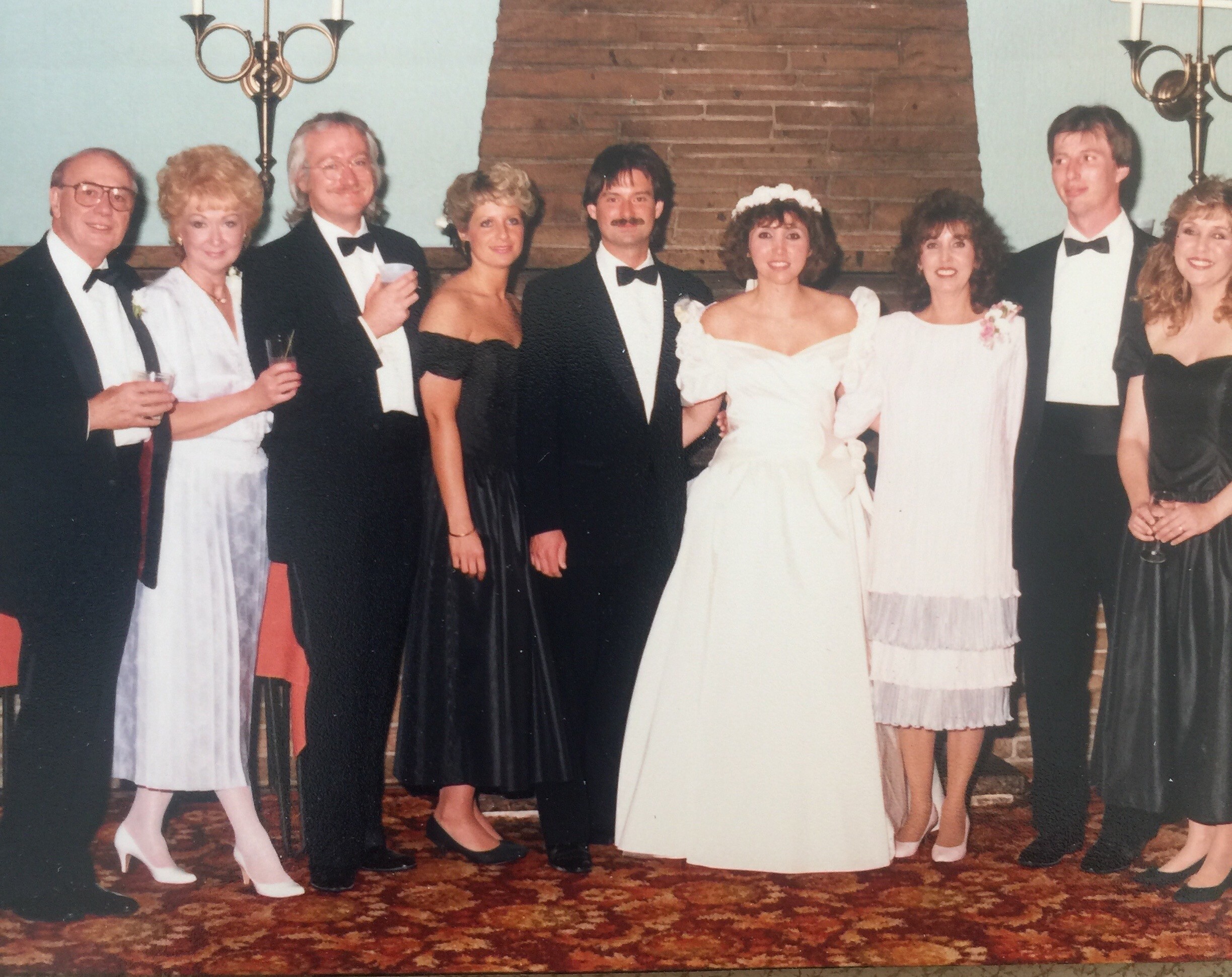 Group of formally dressed people at a wedding, with the bride in a white gown center-right, posing indoors.