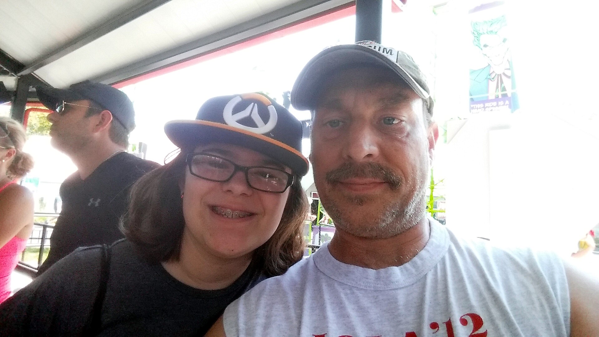 Smiling man and woman take a selfie on a sunny amusement ride, with other riders in the background on a covered platform.