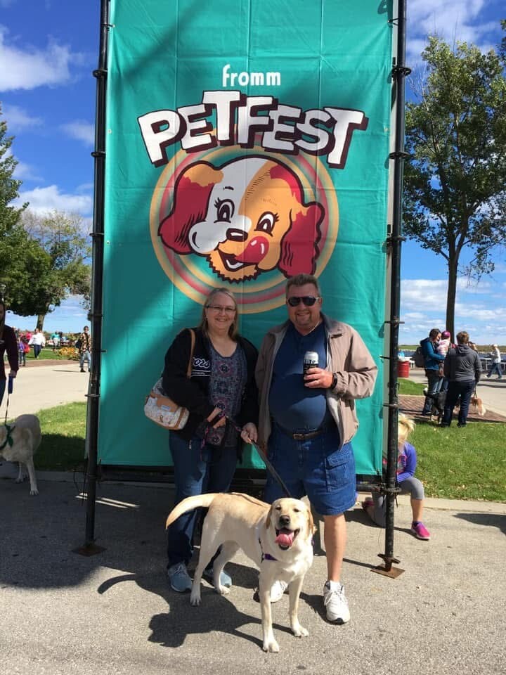 Couple posing with their dog in front of a large teal Pet Fest banner at an outdoor event.