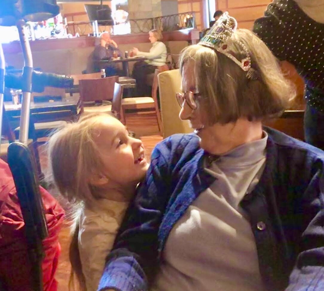Grandmother wearing a birthday crown smiles at a young girl in a restaurant.