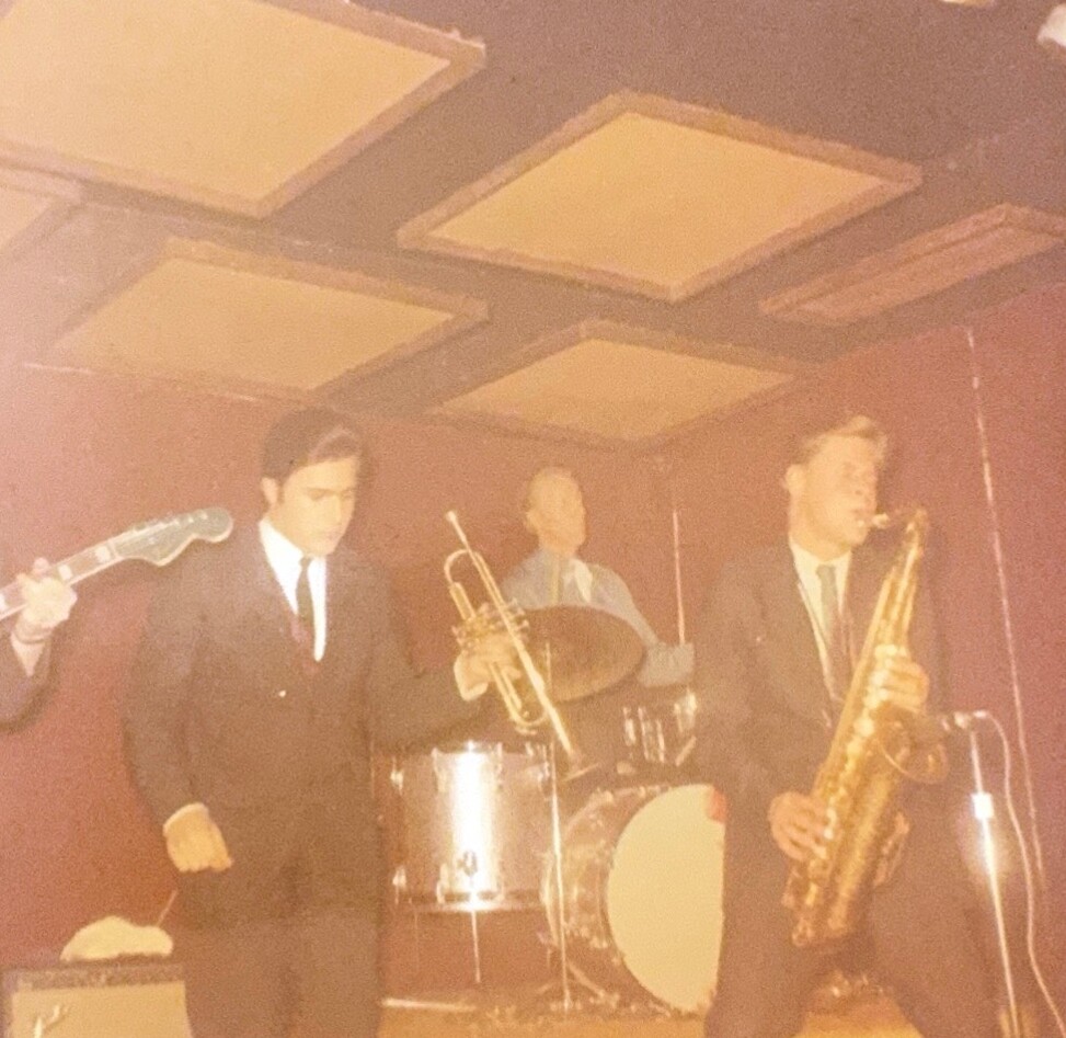 Vintage photo of a four-member band in suits performing on a small stage with guitar, trumpet, drums, and saxophone.
