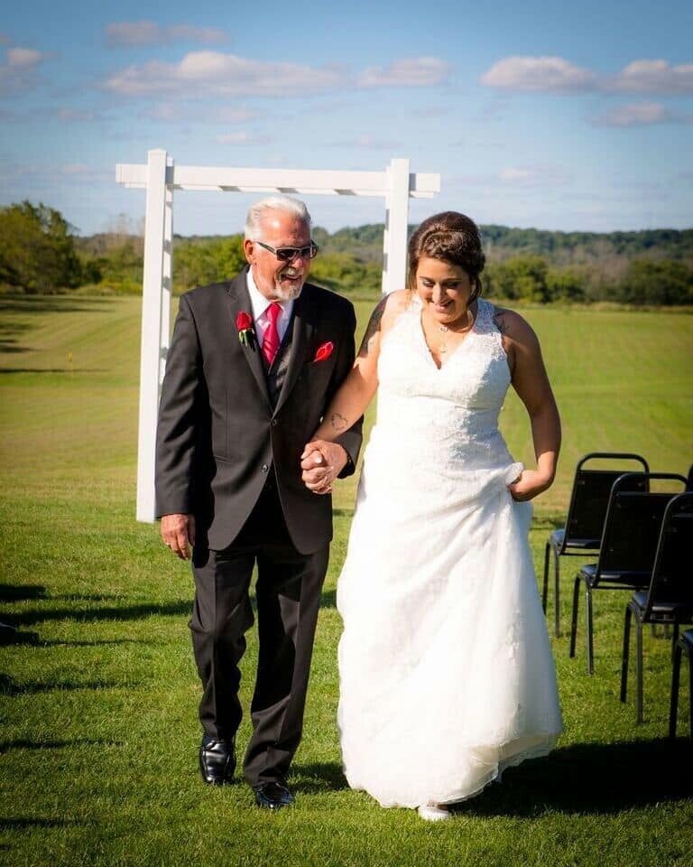 Bride in a white wedding gown walking arm-in-arm with an older man in a black suit and red tie, outdoors on a sunny day near a white wedding arch on green grass.
