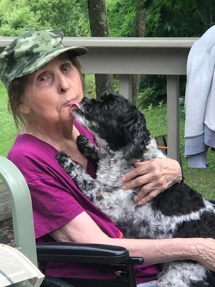 Older woman in a camo hat sits in a chair on a deck as a small black-and-white dog licks her face.