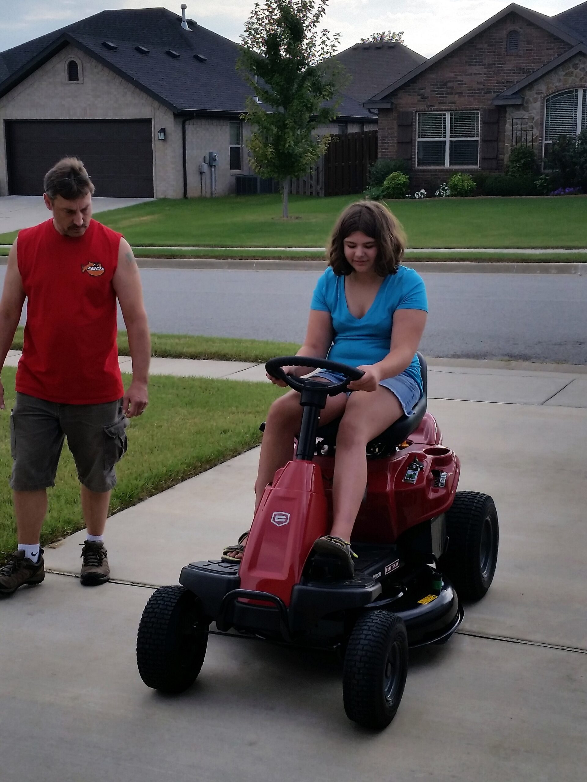 Girl in a blue shirt rides a red riding lawn mower on a driveway while a man in a red sleeveless shirt stands nearby in a suburban neighborhood.
