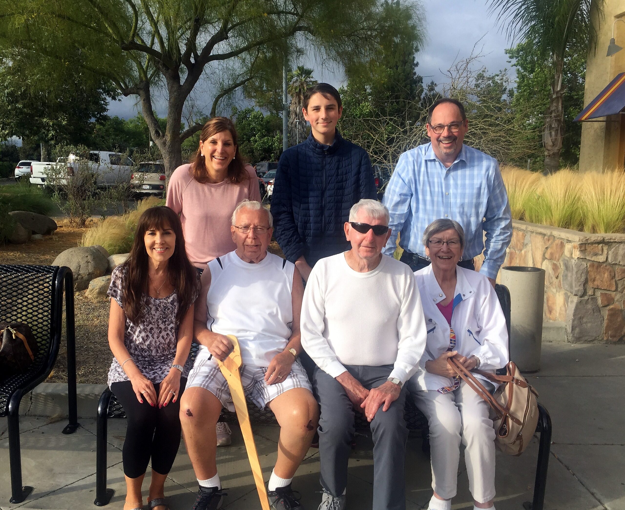 Group of smiling adults posing for a photo outdoors, some seated on a bench and others standing behind a stone wall with trees in the background.
