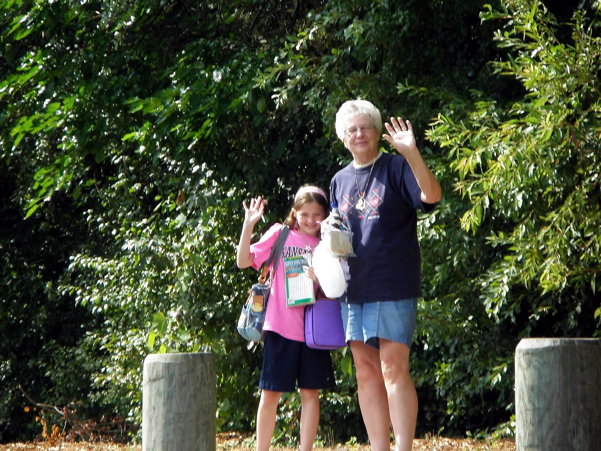 Two people, a woman and a girl, stand outdoors behind a hedge, waving at the camera.” “