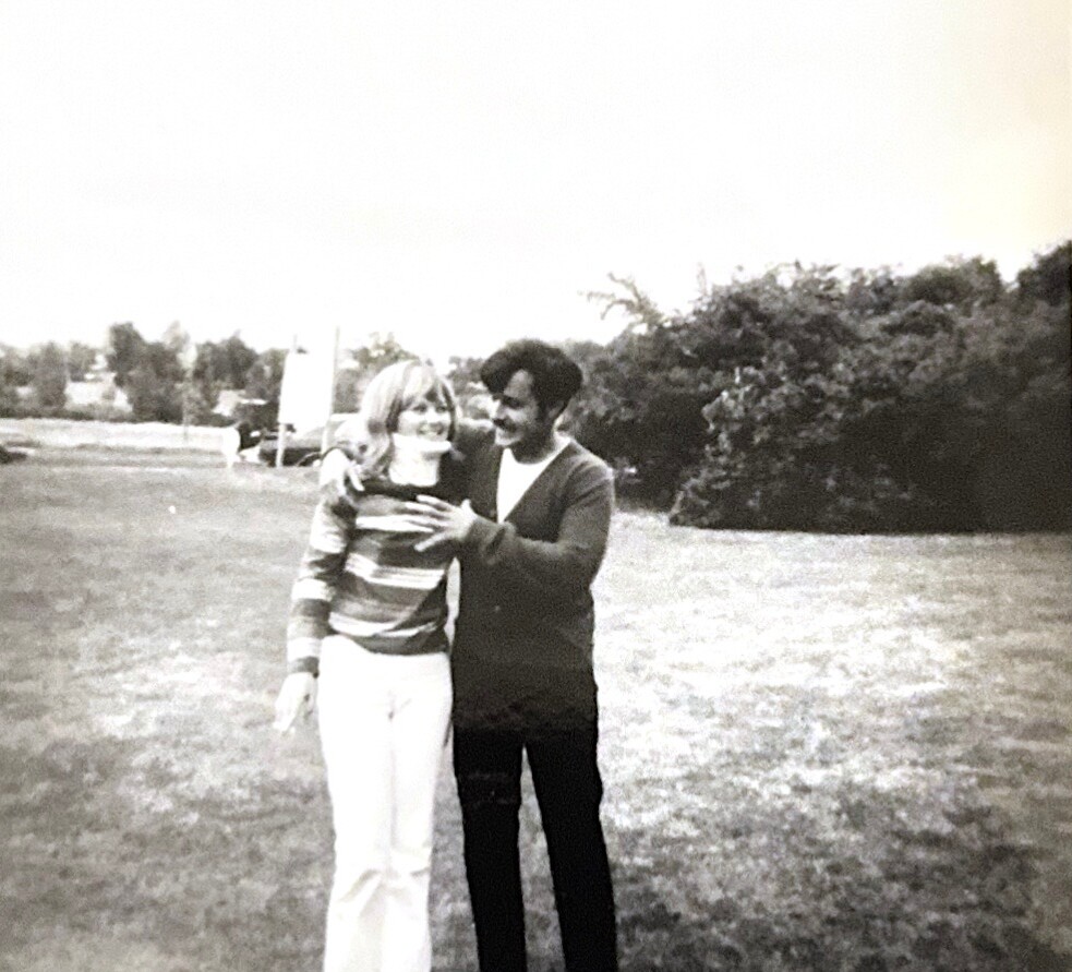 A couple walks together in a park, the man with his arm around the woman as they smile at each other.