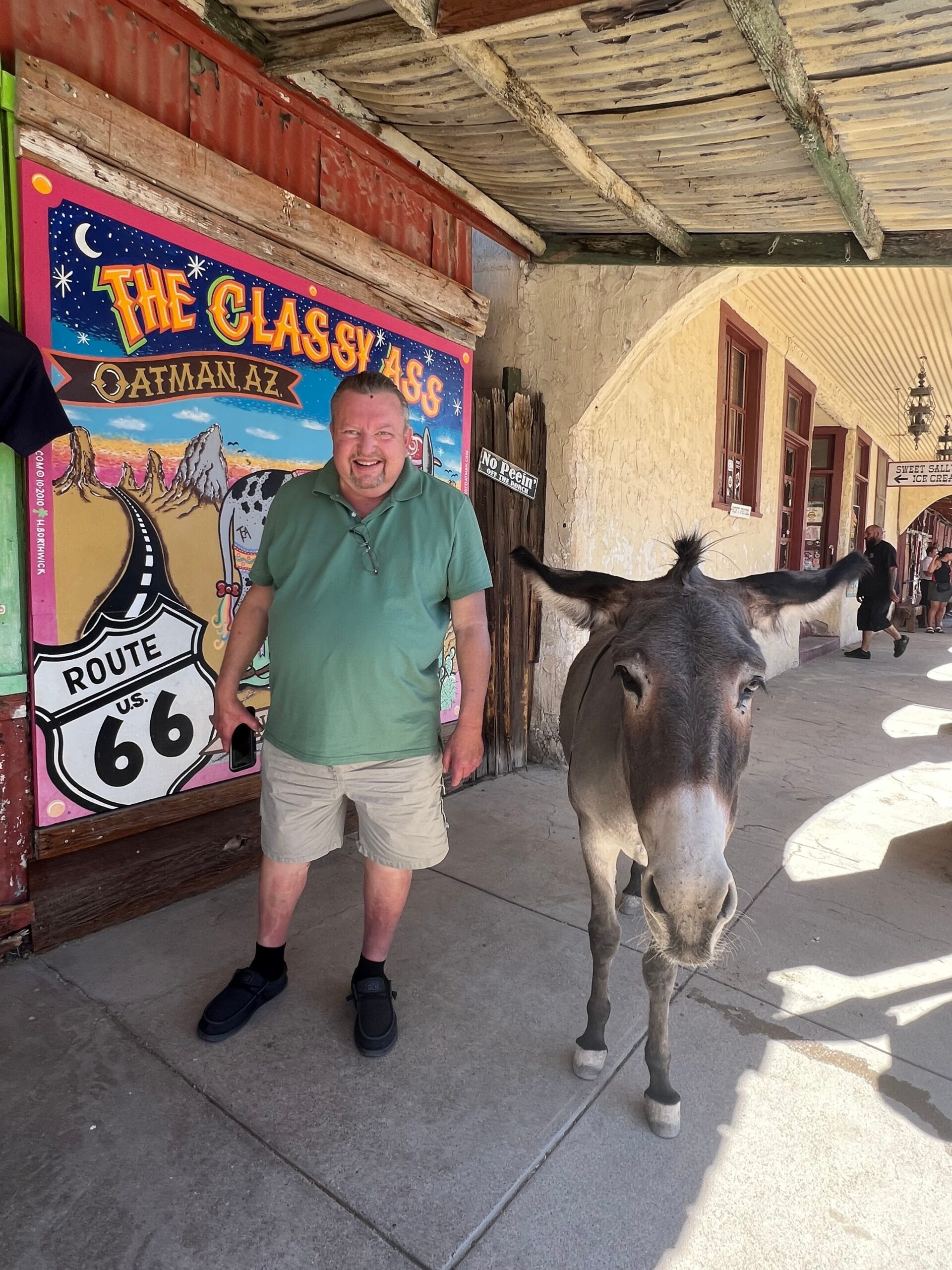 Man in a green polo and khaki shorts stands beside a donkey under a wooden overhang, colorful mural behind them.