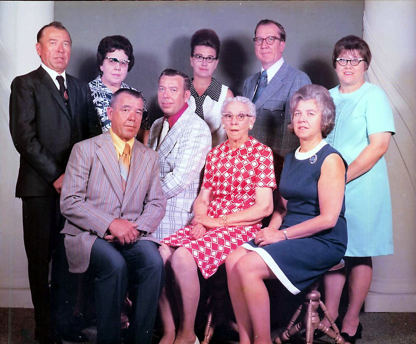 Group of nine adults posing for a formal studio portrait, some seated and others standing, in vintage clothing