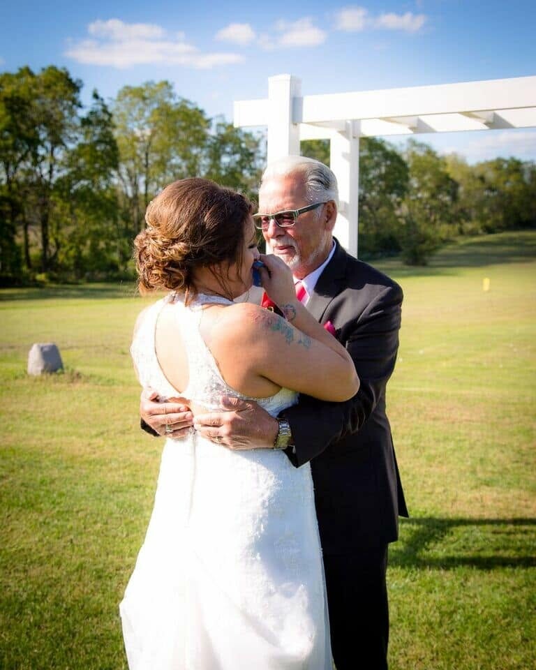 Bride in a white lace dress hugging an older man in a dark suit outdoors on a sunny day.