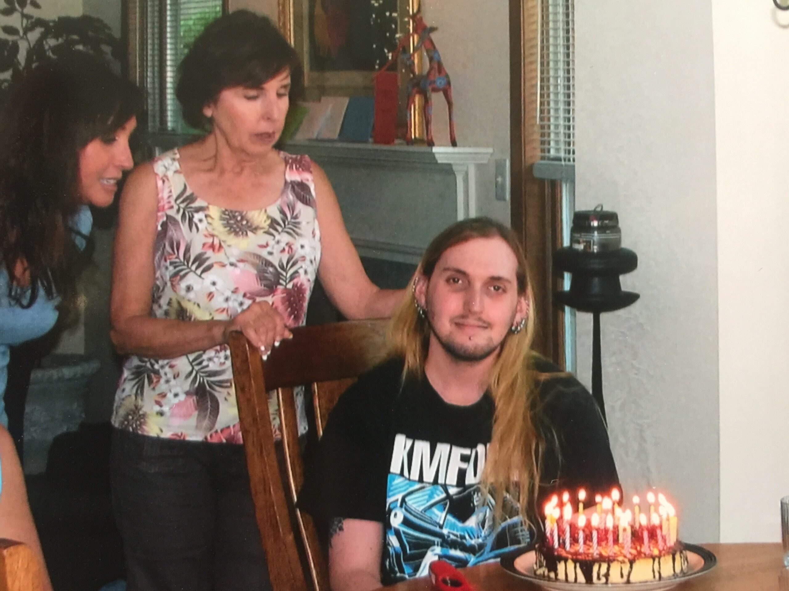 Birthday scene indoors: a woman in a floral dress stands beside a seated man with long blond hair as a cake with lit candles is in front of them.