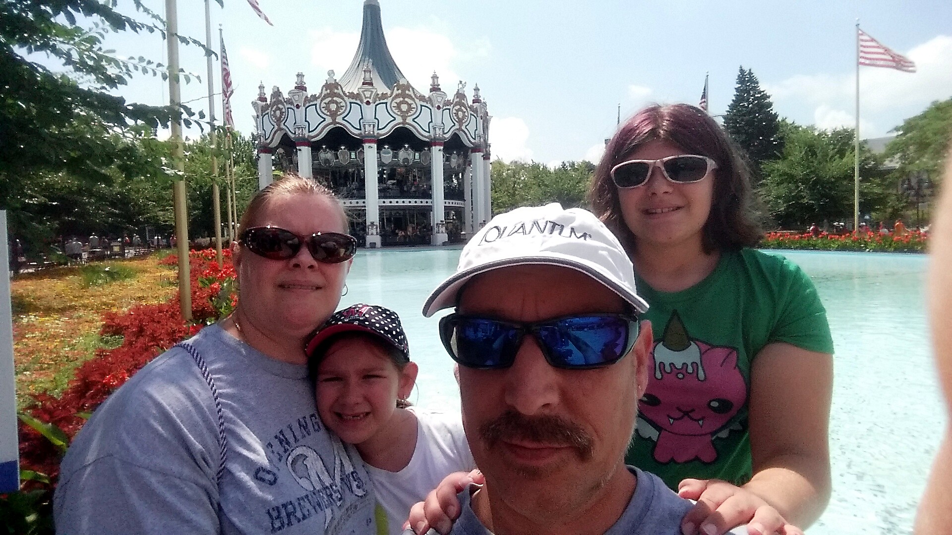 A family of four posing for a selfie in front of a decorative carousel and fountain on a sunny day; man in the foreground wears a white cap and sunglasses, two women and a child stand beside him.