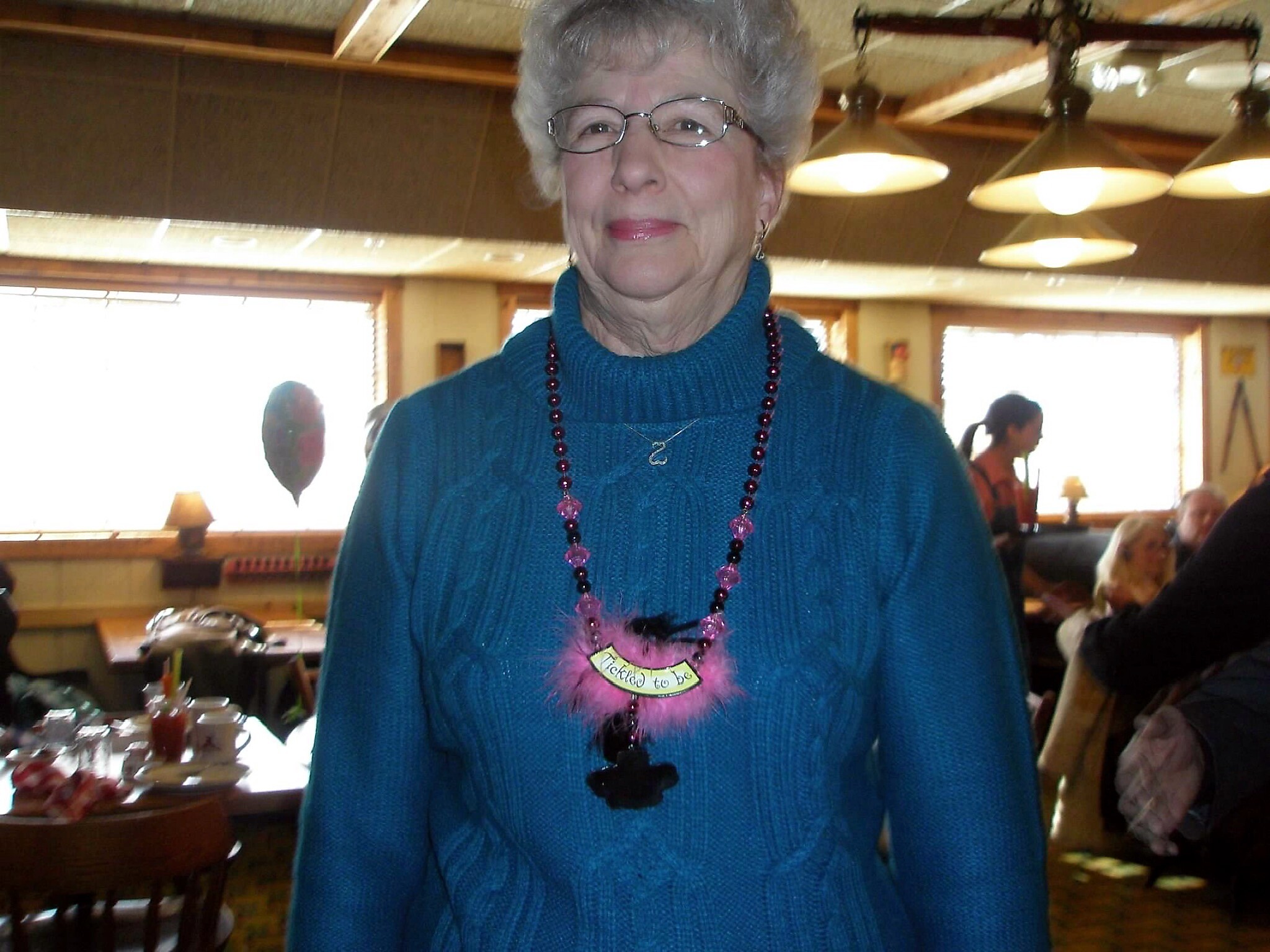 Older woman with short gray hair and glasses, wearing a teal sweater and pink beaded necklace, in a restaurant.