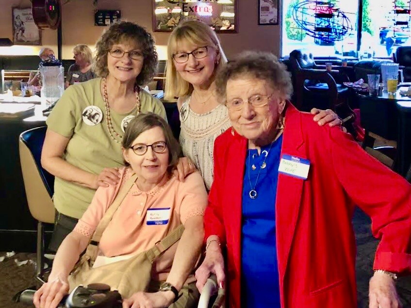 Four smiling women pose for a group photo in a casual restaurant; two stand behind, one sits in a wheelchair, all wearing name tags.