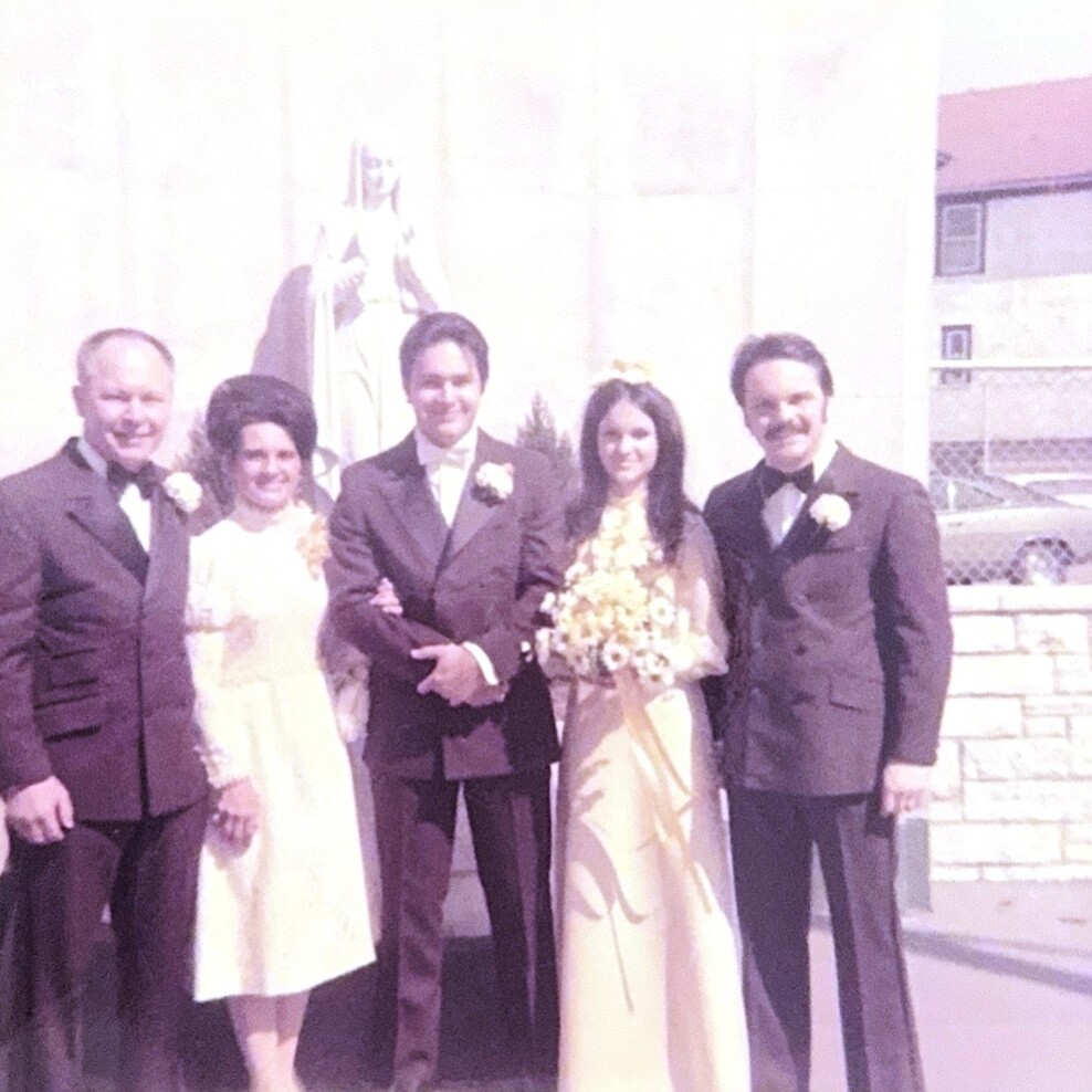 Five adults pose outdoors at a wedding: bride in a light gown holding a bouquet, flanked by grooms and a bridesmaid in white dresses.