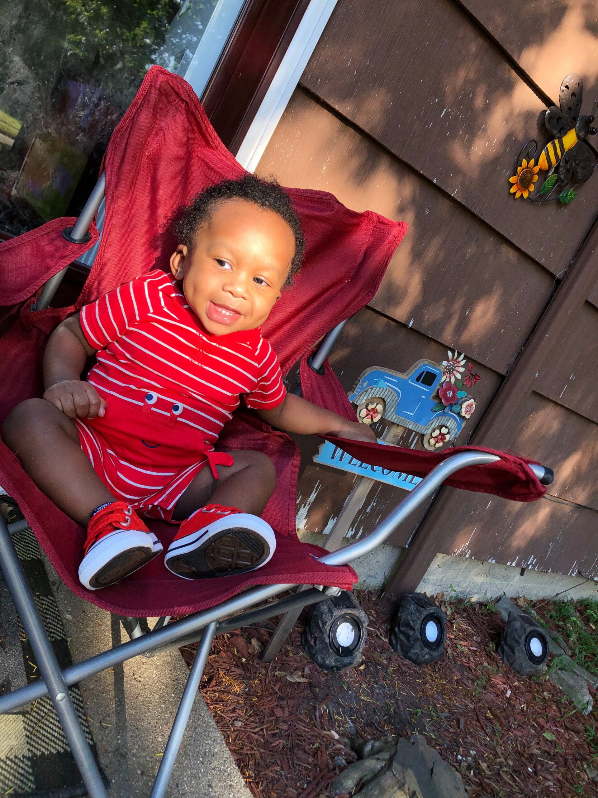 toddler in a red striped outfit sitting in a red stroller outdoors, sunlit near a wooden fence with outdoor decor including a sunflower ornament.