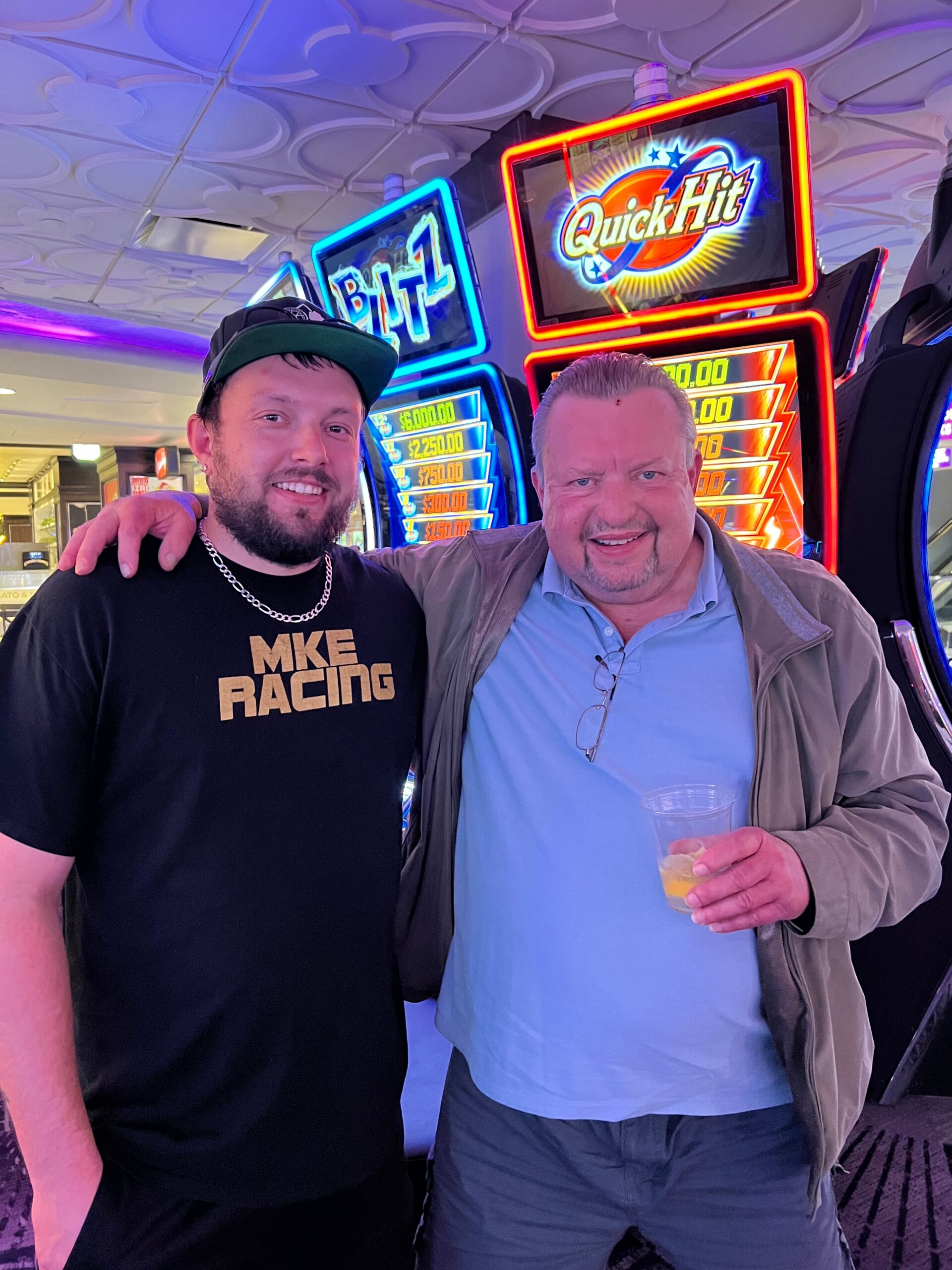 Two men pose together in a casino lobby with bright Quick Hit slot machines in the background; one wears a black shirt and cap, the other a light blue shirt and jacket, holding a glass.