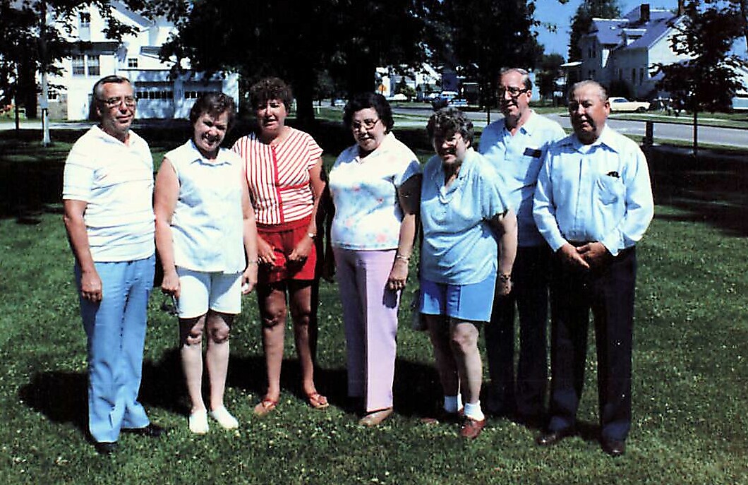 Seven adults stand together on a sunny, grassy lawn in a residential park, posing for a casual group photo.
