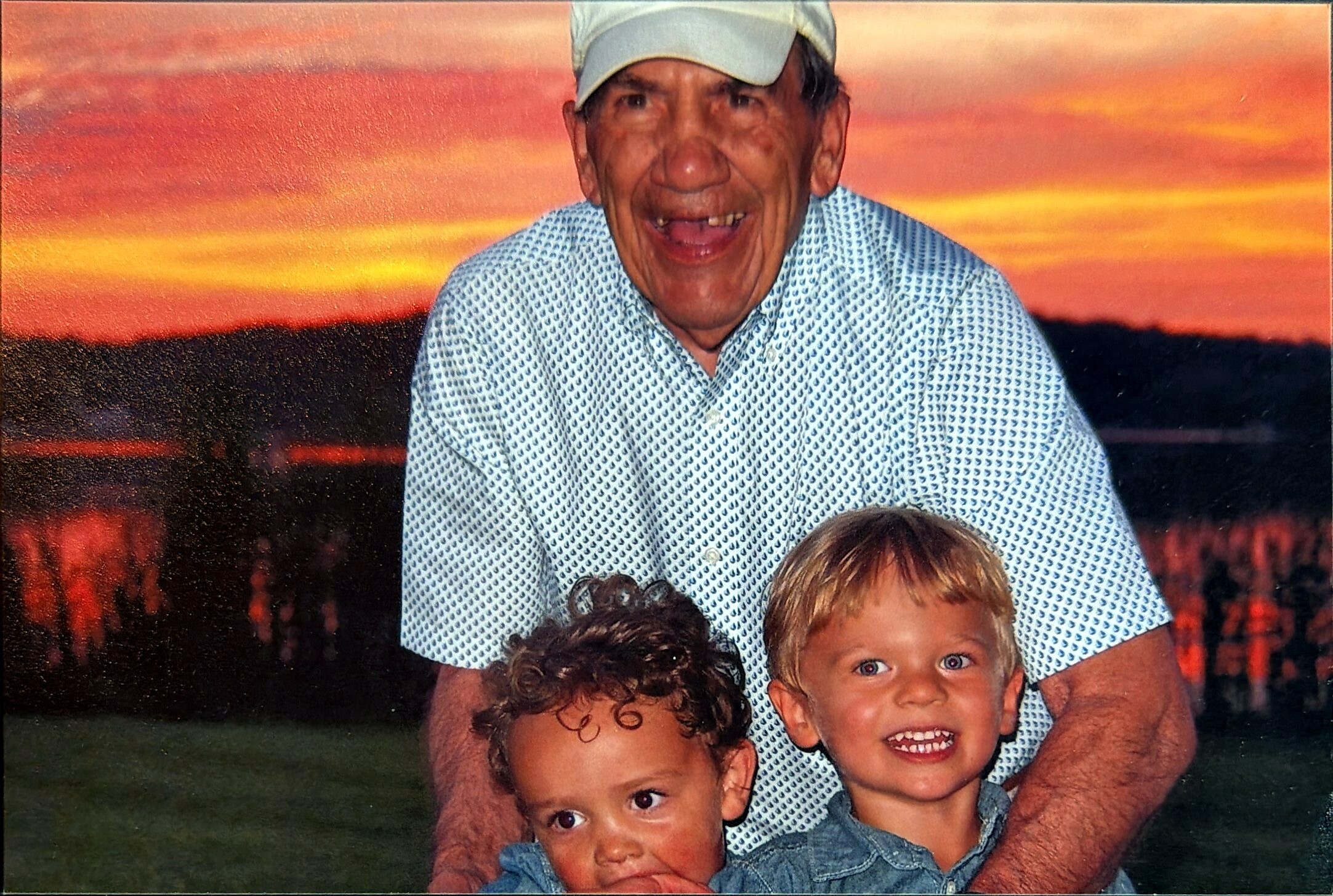 Smiling man in a light cap and patterned shirt posing outdoors with two young children at sunset.